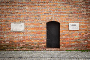 Zdjęcie. Wejście do barbakanu. Obok drewnianych drzwi tablica pamiątkowa. Mur z czerwonej cegły.